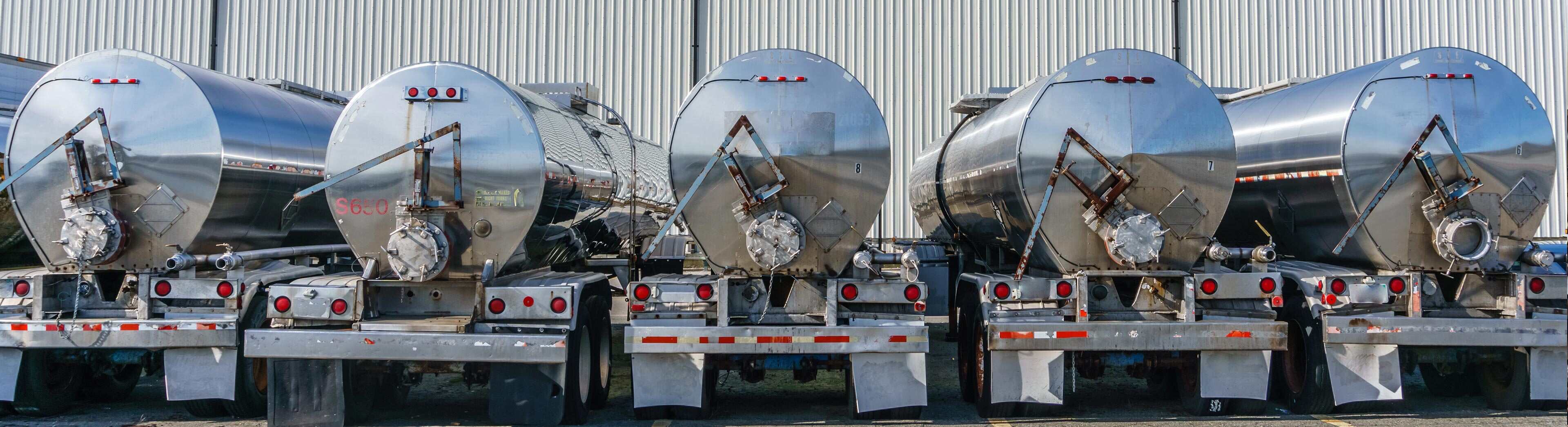 A lineup of massive metal tank trucks used for transporting oil and gas parked in front of a building.