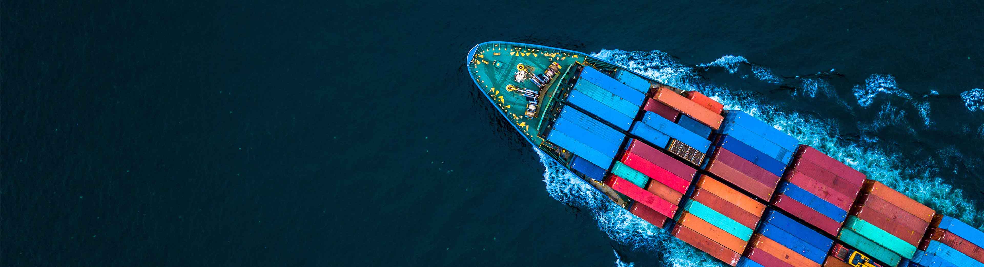 Aerial view of a container ship carrying colourful shipping containers across open water, leaving a wake behind as it travels along a global trade route