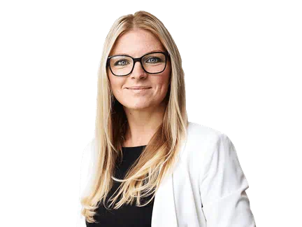 Femme professionnelle aux longs cheveux blonds et portant des lunettes, habillée d’un veston blanc sur un haut noir, se tient debout et sourit avec confiance sur un fond blanc
