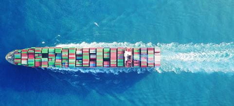 Aerial view of a large container ship sailing through turquoise ocean waters, carrying rows of multicoloured shipping containers.