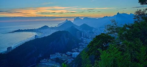 Panoramic view of a coastal city at sunrise, with mountains, a curved shoreline and buildings along the waterfront