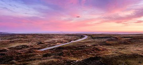 DSV truck driving through an open coastal landscape at sunset, with an aircraft flying across the sky above
