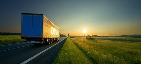DSV:n rekka, jossa on sininen perävaunu ja valkoinen DSV:n logo, ajaa maantiellä peltojen keskellä kohti laskevaa aurinkoa.