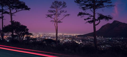 Evening view of a cityscape from a hillside road with silhouettes of trees and a light trail from a passing vehicle under a twilight sky.