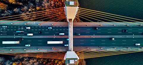 Toma desde arriba de un puente vial de colores, por donde pasan autos y tráileres. En el fondo se ve un cuerpo de agua y un bosque otoñal.