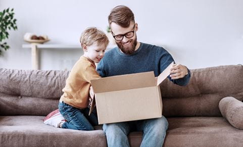 Cheerful bearded man in glasses and curious little boy sitting on sofa and looking inside carton box in cozy living room at home