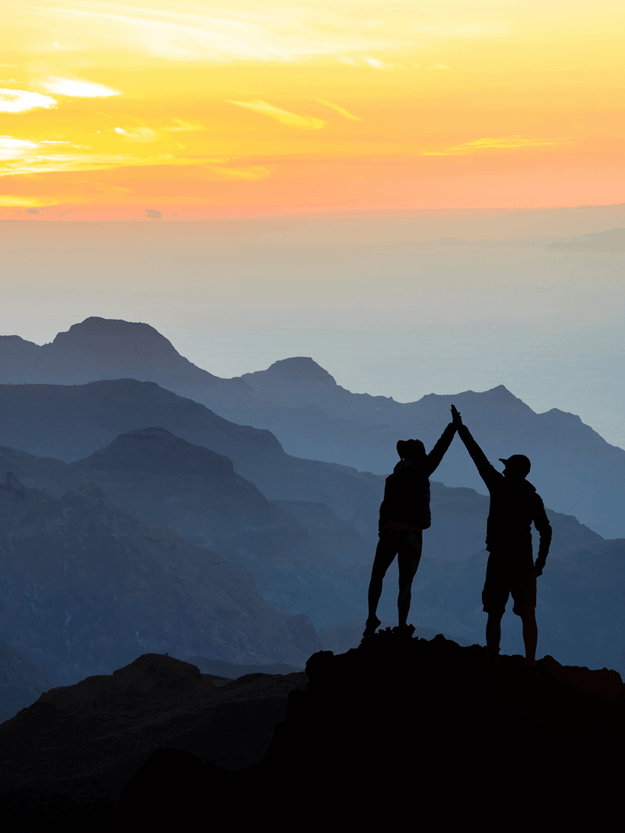 Two persons giving high five to each other on a hill with a sunset behind