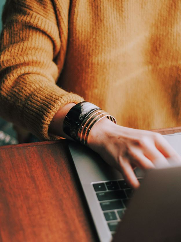 Close-up of a person in a brown sweater using a laptop at a wooden desk, with focus on their hand and a metallic bracelet on their wrist