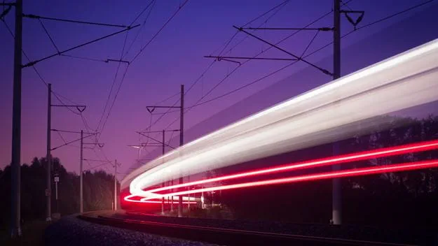 Long-exposure shot of a high-speed train at twilight, showing streaks of red and white lights along curved railway tracks with overhead power lines.
