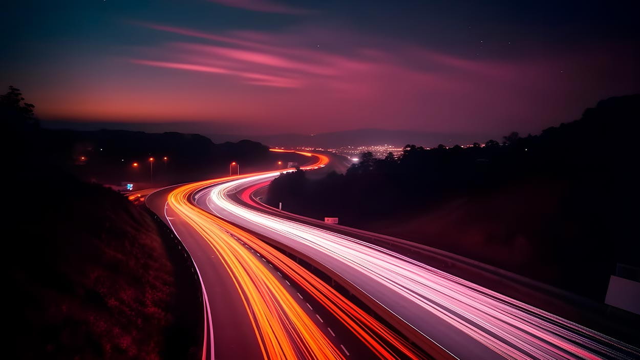 Long-exposure view of highway traffic at night, showing red and white light trails curving through a dark landscape under a colorful evening sky.