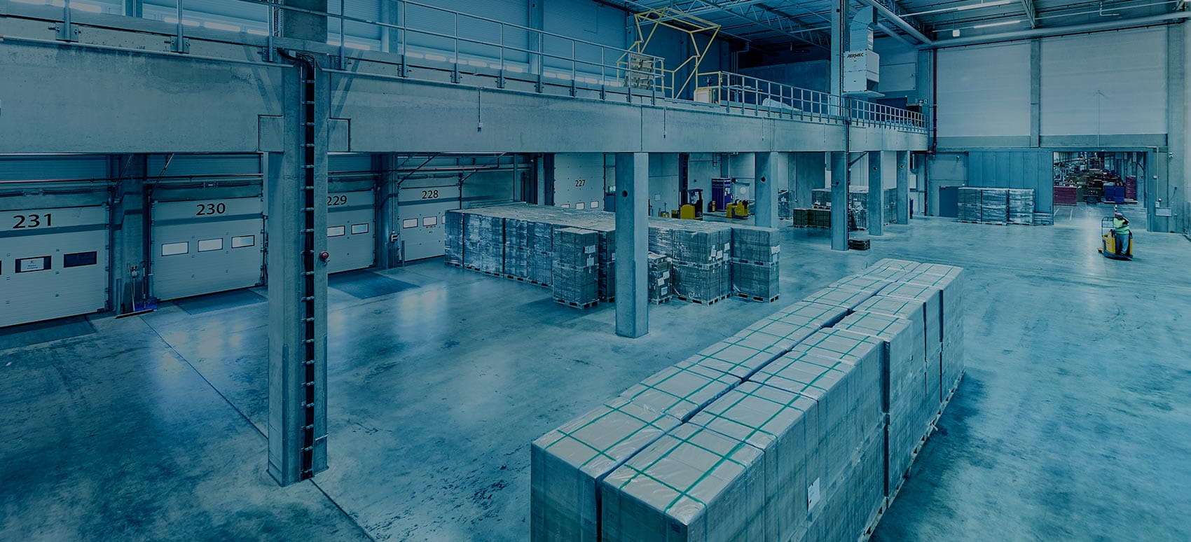 Large warehouse distribution center with loading docks, palletized goods, and workers operating equipment in a spacious storage and logistics facility.