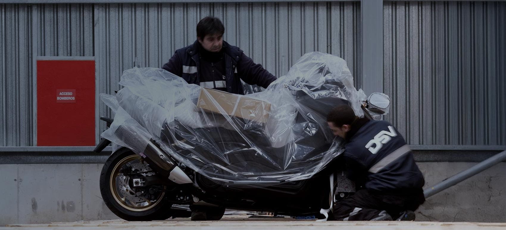  Two DSV workers are handling a large, plastic-wrapped motorcycle on a loading dock.