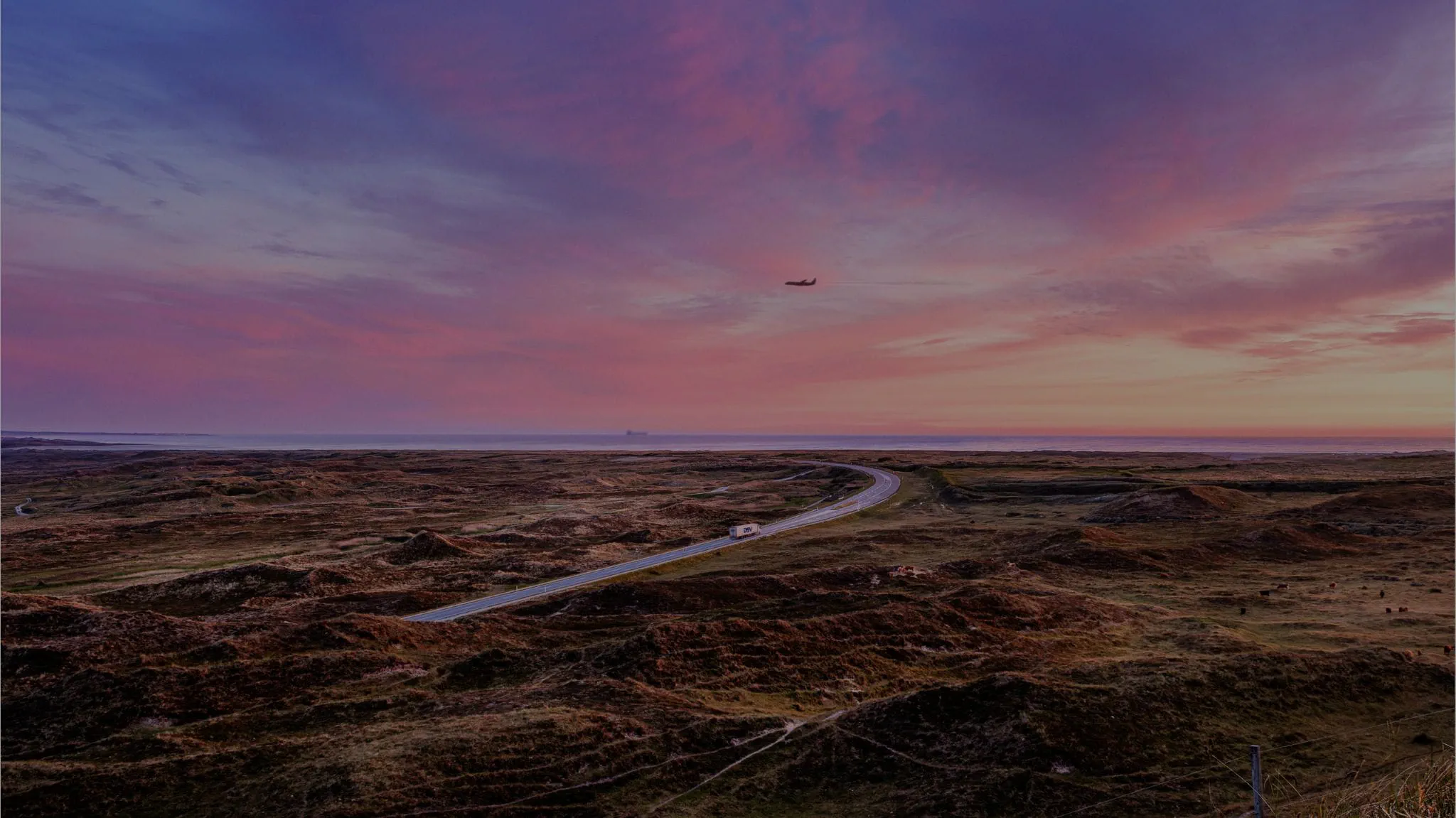 A winding road curves through a rugged, brown coastal landscape under a vivid pink and purple sunset sky, with a small airplane visible in the distance.