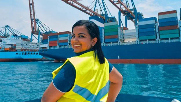 Smiling port worker in a high-visibility vest standing in front of a container ship at a busy harbour