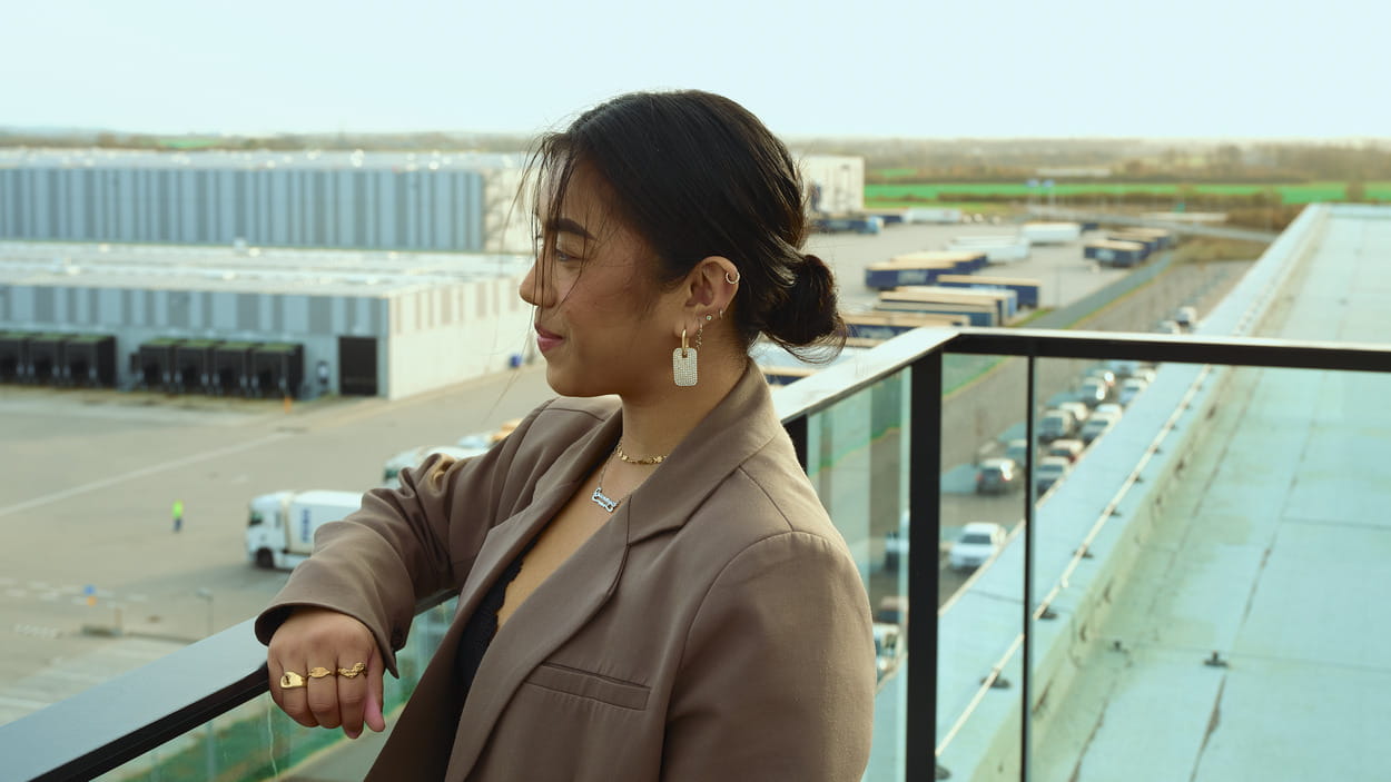  Une femme portant une veste marron contemple une installation logistique avec des véhicules garés.