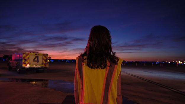 A woman wearing a reflective safety vest stands on an airport tarmac at dusk, looking toward the horizon