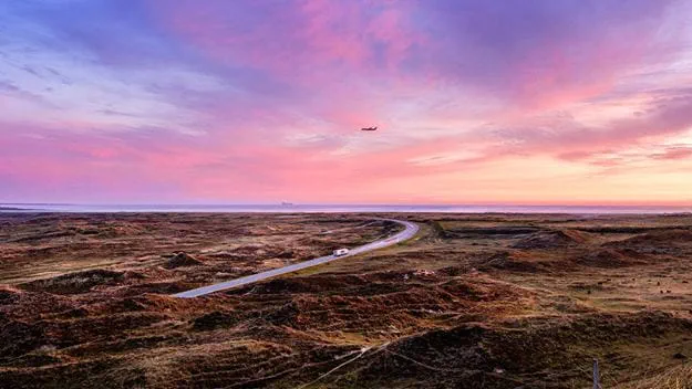 DSV truck driving through an open coastal landscape at sunset, with an aircraft flying across the sky above.