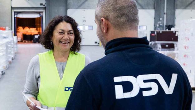 Zwei DSV-Mitarbeiter – eine Frau mit gelber Sicherheitsweste und ein Mann im blauen Hemd – im Dialog in einer Lagerhalle