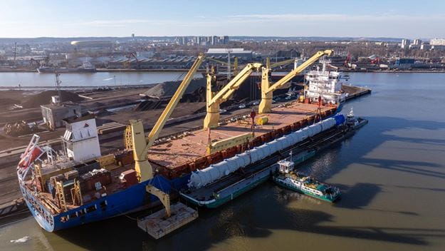 Aerial view of a heavy-lift cargo operation at a commercial port, where a large industrial process vessel is being transferred from a barge to a multipurpose cargo ship using onboard cranes and specialised project logistics equipment