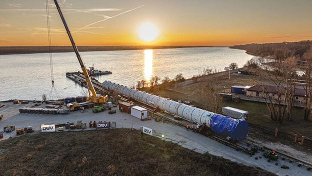 Aerial view of a project cargo operation at sunset, with a large industrial vessel being transferred between rail transport and a barge using a heavy-lift crane beside a riverside terminal