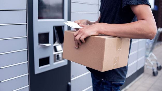A person holding a cardboard parcel and a receipt while using a self-service parcel locker for package collection or delivery