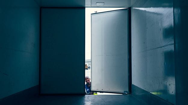 An open door in a large, empty container with minimal lighting, showing a glimpse of the outside