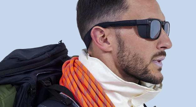 A man in profile wearing dark sunglasses, carrying a large backpack with an orange climbing rope draped over his shoulder. He has short hair and a beard, and he is dressed in a light-coloured, high-collared jacket against a light blue background.