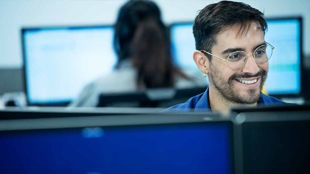 Office employee smiling while working at a computer, with colleagues in the background