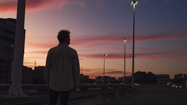 A person standing on an overpass at sunset, looking towards a city skyline and evening traffic
