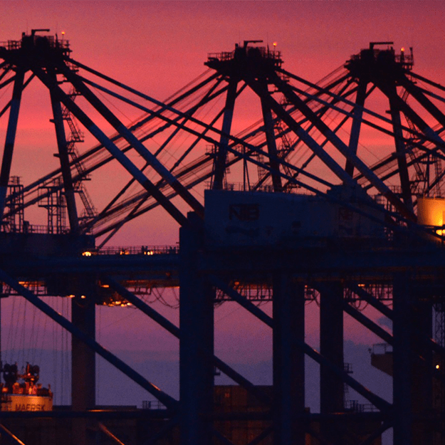 Silhouetted cargo cranes against a vibrant pink and purple sunset sky at a port