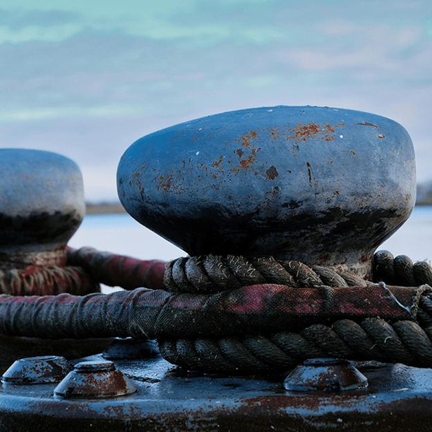 A close-up view of two sturdy metal bollards on a dock, securing thick, weathered ropes against a backdrop of calm waters and a distant shoreline