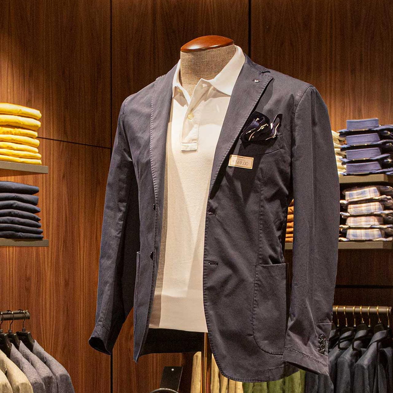 A mannequin dressed in a stylish navy blazer with a pocket square and a white polo shirt, displayed against a warm wooden background