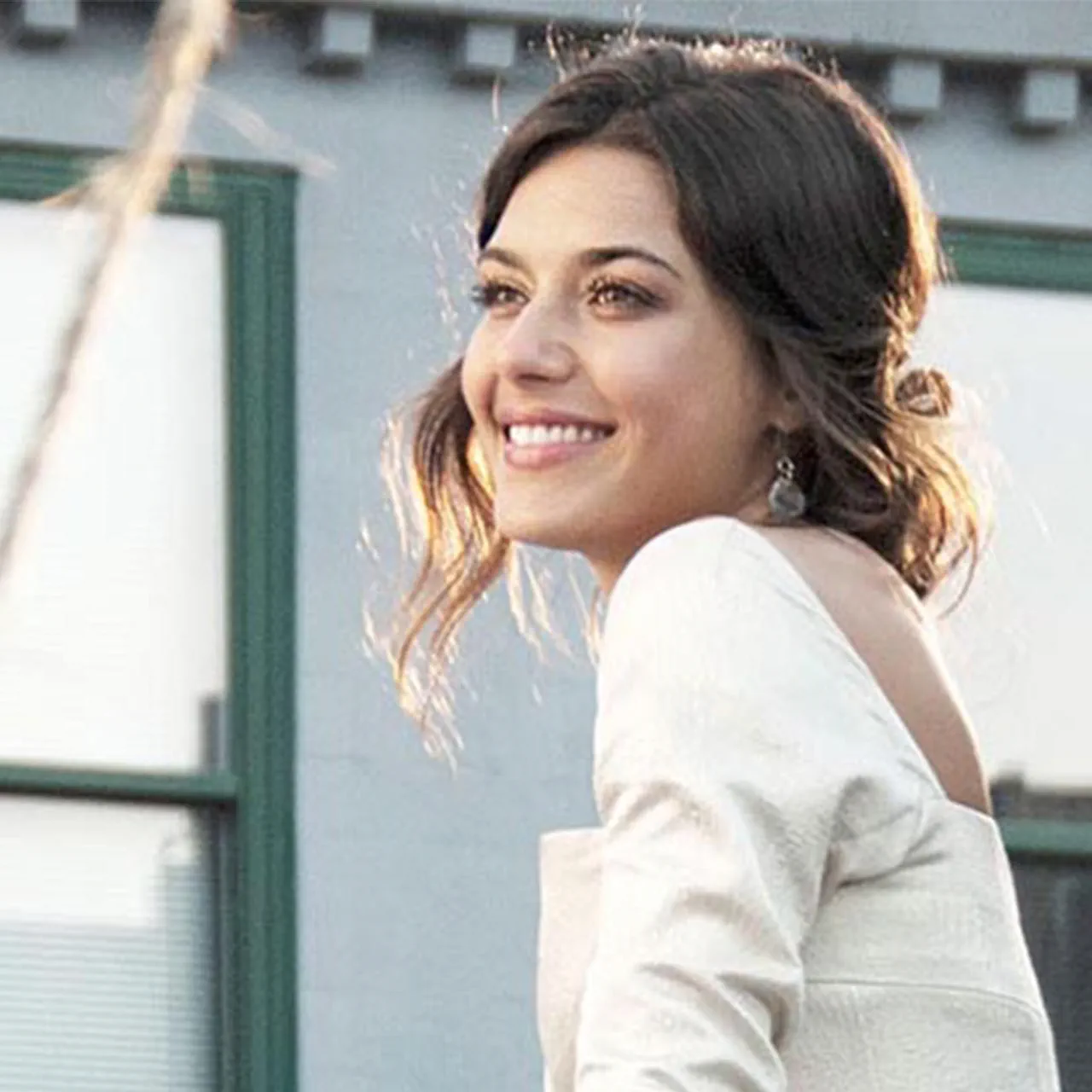 A smiling woman with styled hair, wearing a light-coloured outfit and delicate earrings, captured in soft, natural lighting