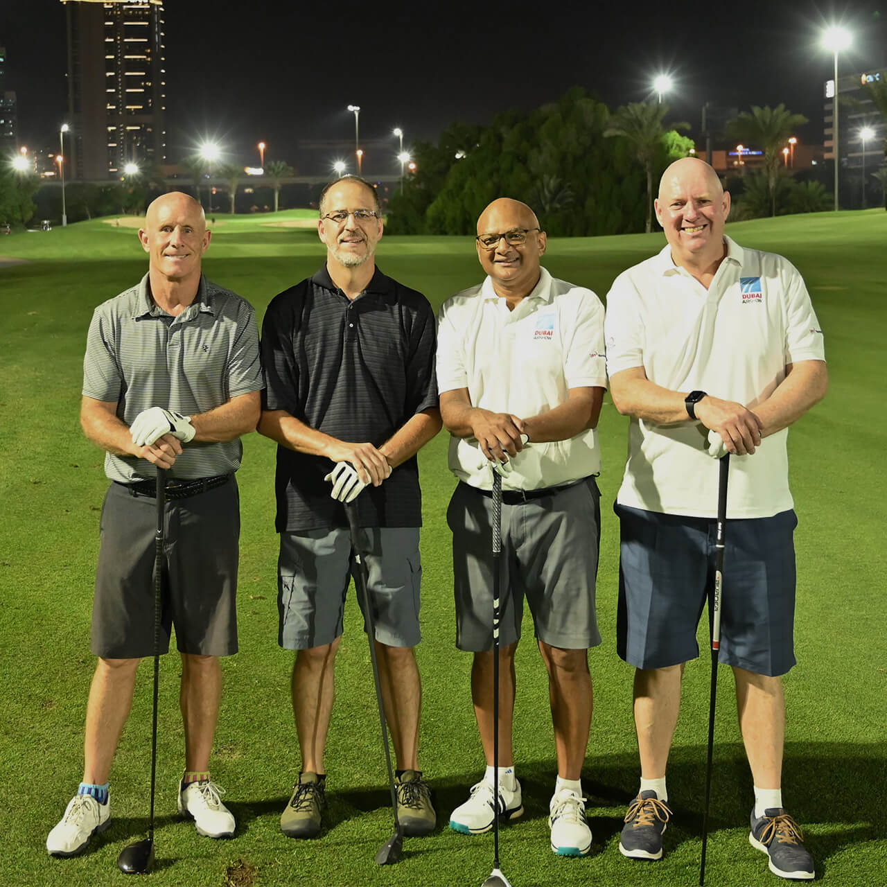 Four men stand on a lit golf course at night, smiling and holding golf clubs. The background features trees and city lights. They appear relaxed and cheerful.