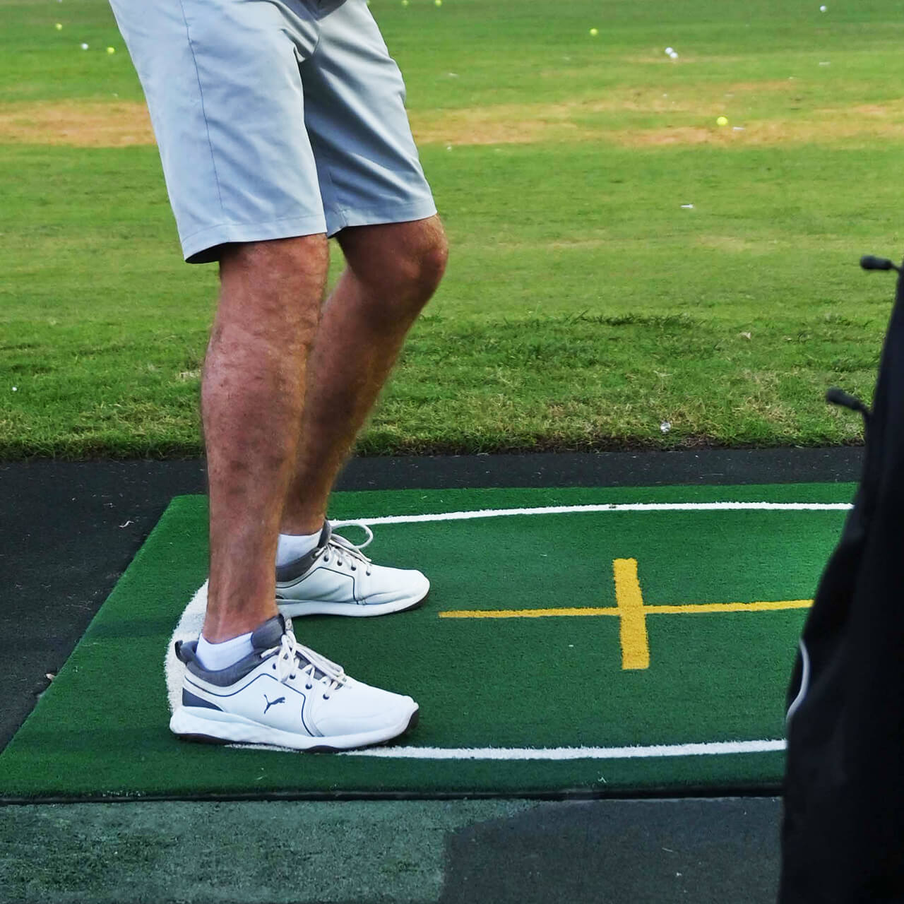 Close-up of a person wearing white sneakers, stepping onto a green grass mat marked with a yellow cross. The tone is casual and focused.