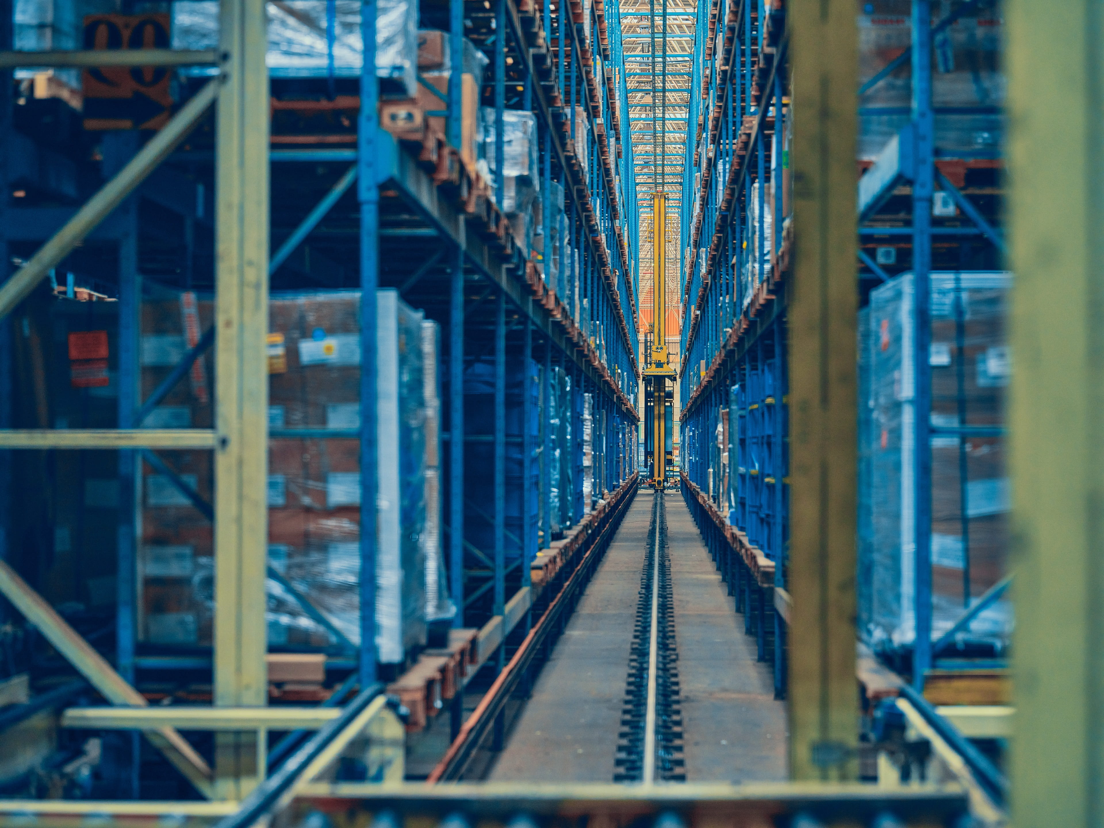 A warehouse aisle with tall blue shelves stacked with pallets and a yellow automated machine on the center track.