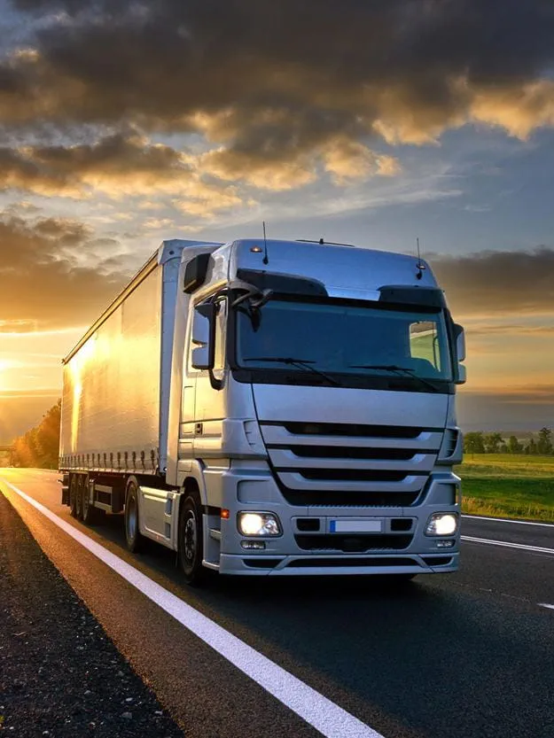 A large freight truck driving on a country road at sunset, with dramatic clouds and golden sunlight casting reflections on the vehicle and surrounding landscape