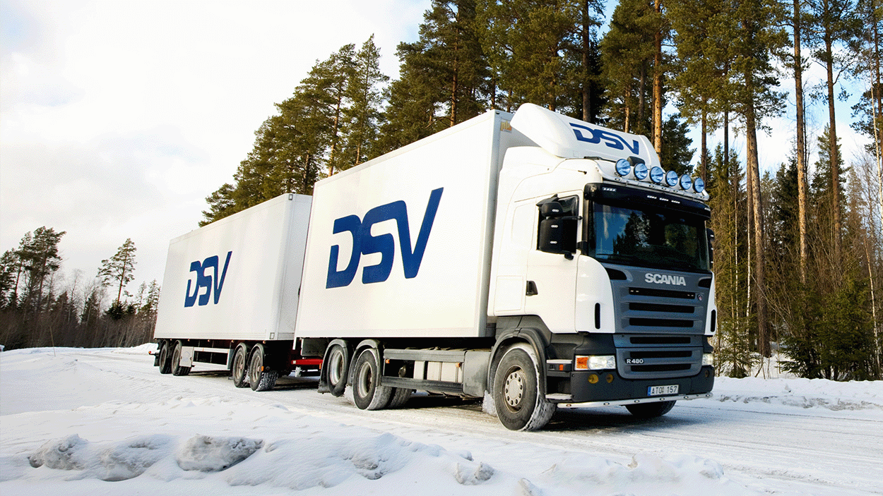 Un camion articolato DSV bianco con due rimorchi che attraversa un paesaggio boschivo innevato, con alti pini che costeggiano la strada