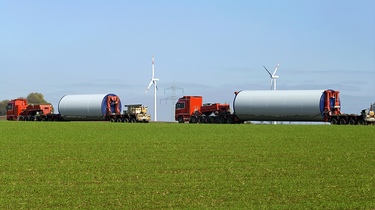 Due camion rossi pesanti trasportano sezioni cilindriche di torri eoliche di grandi dimensioni su rimorchi a pianale ribassato attraverso un campo erboso, con turbine eoliche e linee elettriche sullo sfondo
