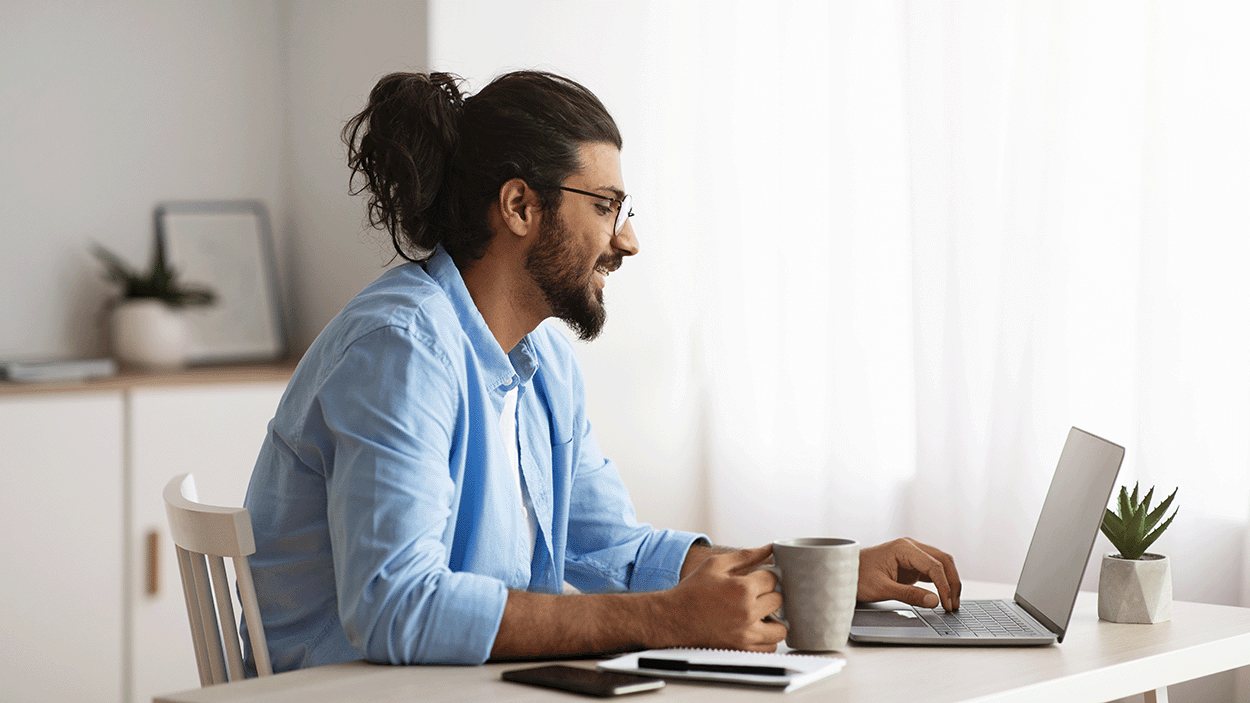 Una persona seduta alla scrivania, sorridente mentre lavora al computer portatile e tiene in mano una tazza, in un luminoso ufficio domestico.